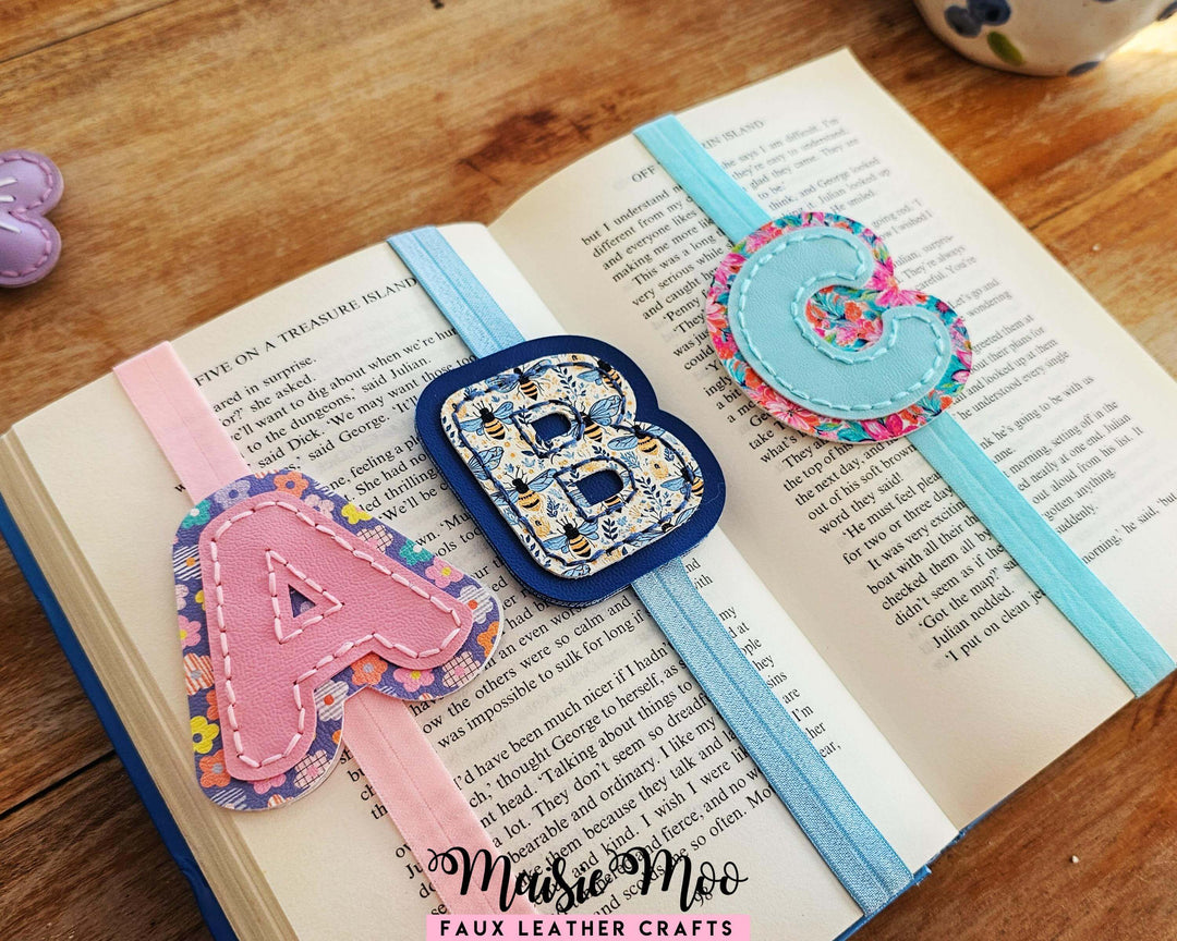 Alphabet Bookmark SVG Set featuring faux leather bookmarks with stitched initials A, B, and C on an open book.